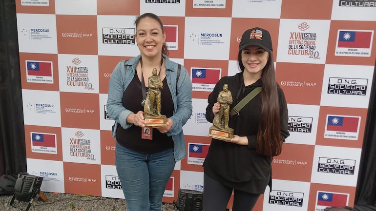 Dos mujeres sonrientes, una con cabello recogido y chaqueta de mezclilla, otra con gorra, sostienen estatuillas en un evento cultural.
