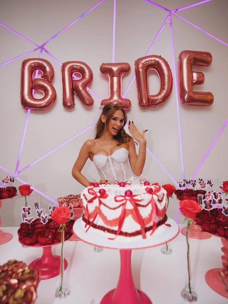 Mujer sonriente con corsé blanco levanta la mano en un salón decorado con globos rosas y pasteles de cumpleaños.