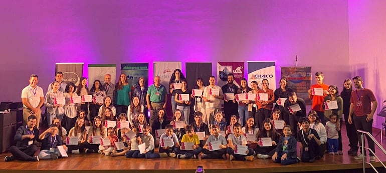 Grupo diverso de jóvenes y niños sonríen sosteniendo certificados, en un ambiente festivo con fondo morado y banners publicitarios.