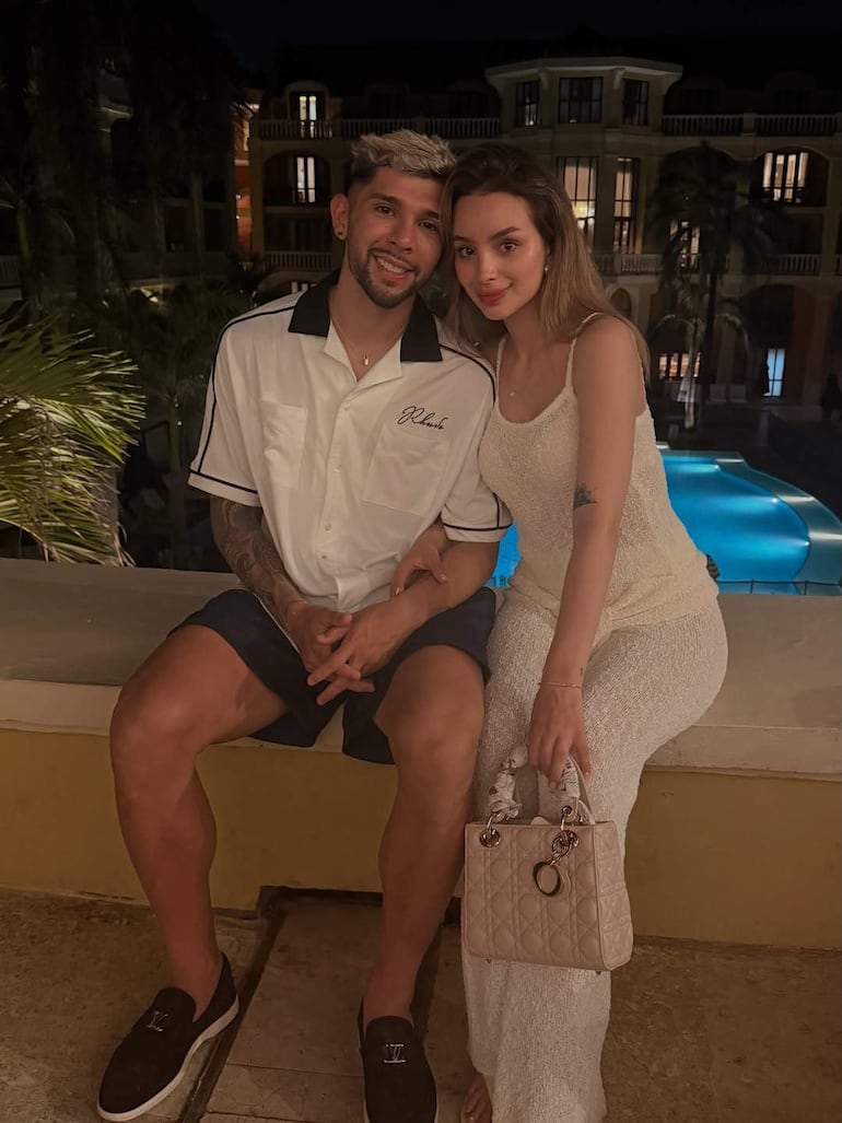 Ramón Sosa con camisa blanca y pantalones cortos, y Julio Enciso con vestido blanco, ambos sonriendo junto a una piscina iluminada.