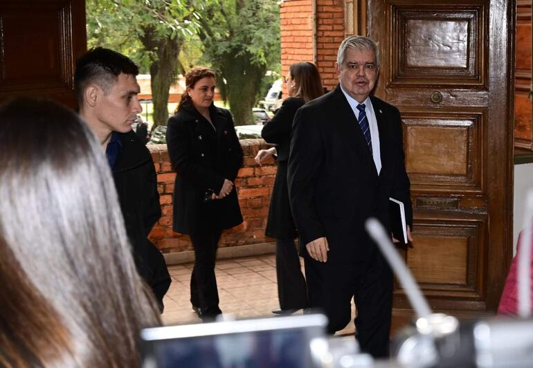 Hombre de cabello gris con traje oscuro y corbata azul habla, mientras un joven en camisa azul escucha atentamente en un ambiente formal.