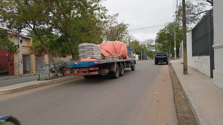 Camión de carga con sacos de cemento cubiertos por una lona naranja en una calle del barrio Trinidad, rodeado de casas y árboles.