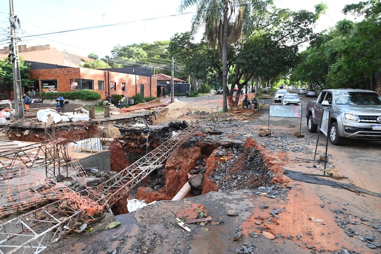 Escena en callejón con excavación y escombros, edificio de ladrillo al fondo y vehículo gris estacionado.