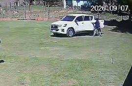 Dos hombres, uno con abrigo oscuro y sombrero, corren hacia una camioneta blanca en un entorno rural.