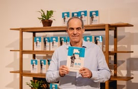 Jorge Mendelzon sostiene su libro 'DE PIE', vistiendo una camisa clara y sonriendo frente a una estantería con más copias.