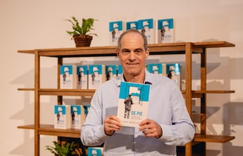 Jorge Mendelzon sostiene su libro 'DE PIE', vistiendo una camisa clara y sonriendo frente a una estantería con más copias.