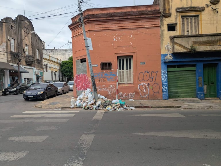 Montículo de basura en la acera frente a edificio naranja con grafitis y vehículos estacionados a la izquierda.