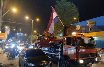 Personas en camioneta roja sosteniendo bandera de Paraguay, mientras celebran en la calle durante la noche, rodeados de vehículos y luces.