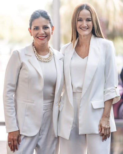 Dos mujeres con trajes blancos sonríen, una con cabello recogido y collar de perlas, en un entorno luminoso y formal.