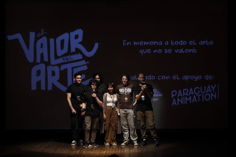 Siete personas en el escenario con vestimenta oscura y una mujer en blusa clara, junto a una pantalla que dice 'El Valor del Arte'.