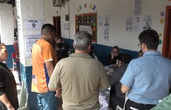 Una mujer con chaqueta negra observa, mientras un hombre de camiseta naranja charla en un salón de votación con mesas identificadas.