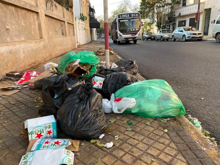 Montones de basura se acumulan en la calle, con un autobús y vehículos estacionados, en un ambiente de descuido urbano.