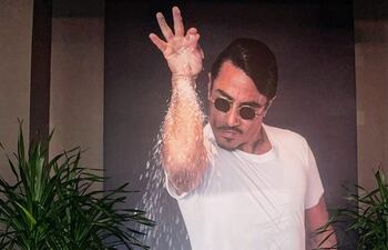 El chef turco Salt Bae posa recostado en un sillón junto a un retrato gigante de él mismo que lo muestra en su característica pose al salar la carne.