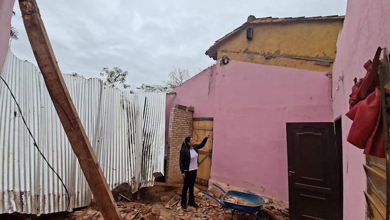Mujer en chaqueta negra señala daño en la pared de una estructura blanca, rodeada de materiales de construcción en un día nublado.