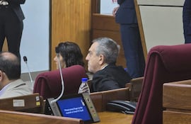 Yamil Esgaib sentado en su banca con expresión tranquila, rodeado de un ambiente formal y atención de otros presentes.