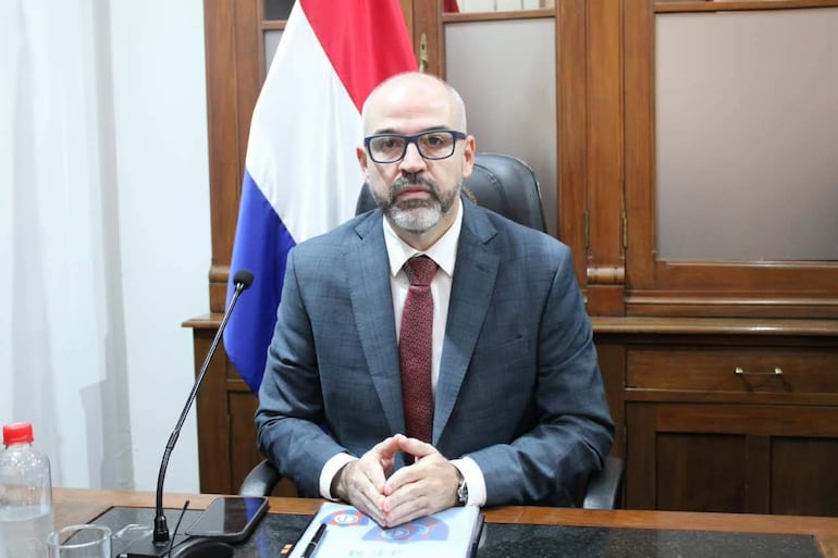 Enrique Javier Berni Brítez sentado en escritorio, con traje gris y corbata burdeos, frente a la bandera de Paraguay.