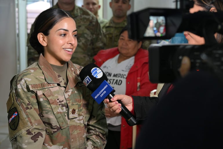 Mujer militar sonriendo mientras es entrevistada, rodeada de hombres en uniformes camuflados en un ambiente interior.