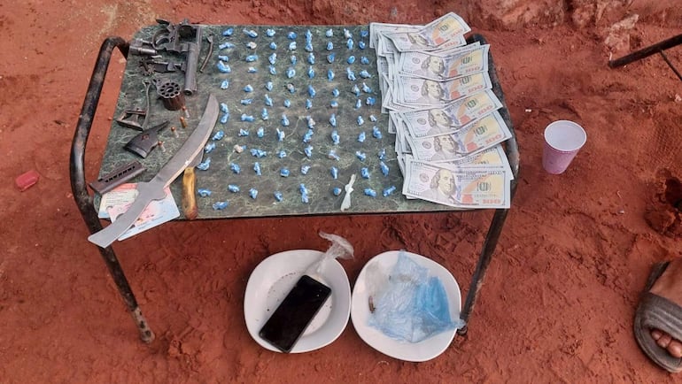 La imagen muestra una mesa de metal con varios objetos sobre ella, situada en un entorno de tierra roja. En la superficie se pueden ver numerosos envoltorios de color azul, que podrían contener sustancias ilícitas, distribuidos de manera ordenada. También hay una cantidad considerable de billetes de cien dólares en un lado de la mesa, junto con un arma de fuego y una especie de machete, lo que sugiere una escena asociada a actividades delictivas.