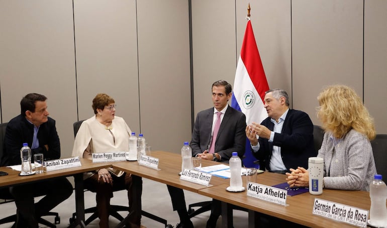Luis Ramírez y Marco Riquelme conversan en mesa con Katja Afheldt y otros, documentos y botellas de agua presentes en sala con bandera nacional.