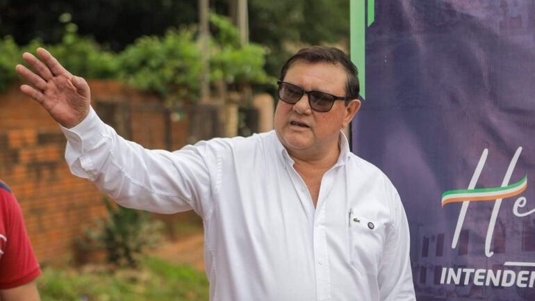 Hombre robusto con camisa blanca gesticula mientras se comunica, rodeado de vegetación y una pared de ladrillos.