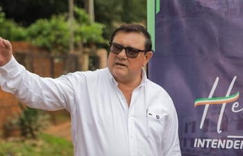 Hombre robusto con camisa blanca gesticula mientras se comunica, rodeado de vegetación y una pared de ladrillos.