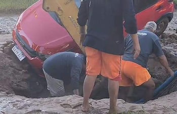 Cuatro hombres en camisetas grises y pantalones cortos naranjas trabajan junto a una retroexcavadora y un auto rojo hundido.