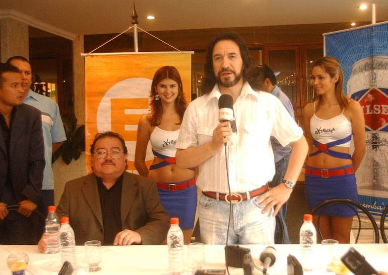Marco Antonio Solís con micrófono en mano, mientras un hombre con gafas lo escucha y dos mujeres con camisetas blancas sonríen a su lado.
