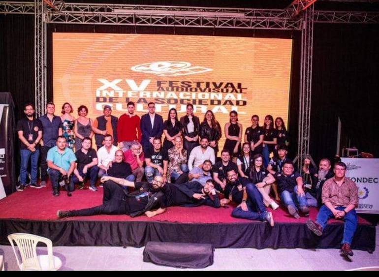 Hombre en traje oscuro en el centro del escenario, rodeado de un grupo sonriente de realizadores durante un festival de cine.