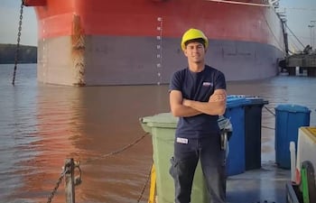 Joven con casco amarillo y camiseta oscura, de pie con brazos cruzados, junto a la embarcación G.W. Fortune en un puerto.