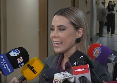 Lizarella Valiente, con cabello lacio y vestimenta formal, habla rodeada de micrófonos en un ambiente institucional.