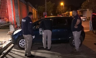 Tres hombres policías con uniformes oscuros inspeccionan un coche azul, uno agachado revisando el interior y los otros observando.
