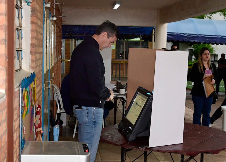 Santiago Peña observa la máquina de votación con papeles en mano, en un ambiente de votación formal con otros votantes detrás.