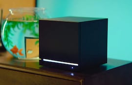 Dispositivo negro en forma de cubo y pecera con goldfish anaranjados, sobre mueble de madera oscura en ambiente luminoso.