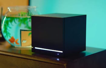 Dispositivo negro en forma de cubo y pecera con goldfish anaranjados, sobre mueble de madera oscura en ambiente luminoso.