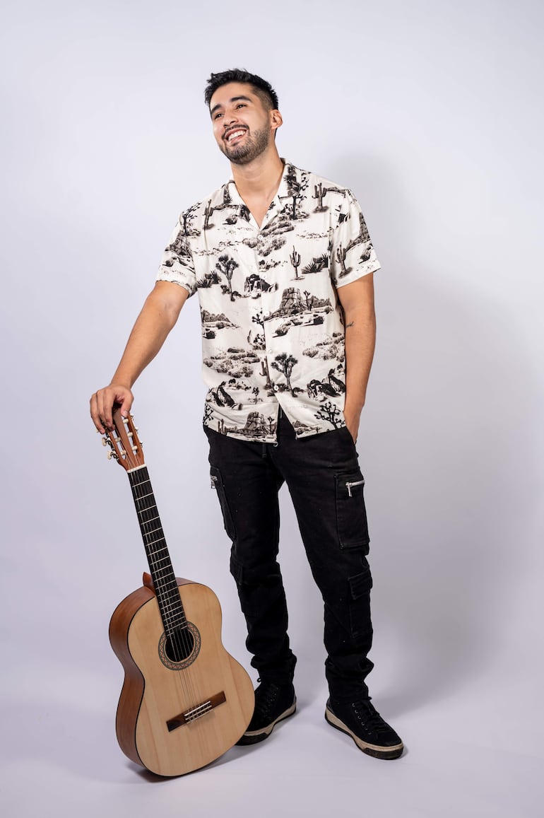Hugo Ferreira de pie, con camisa clara y pantalones oscuros, sonriente junto a su guitarra en fondo neutro.