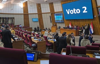 Mujer de pie en podio hablando, rodeada de miembros del legislativo en un recinto con asientos rojos.