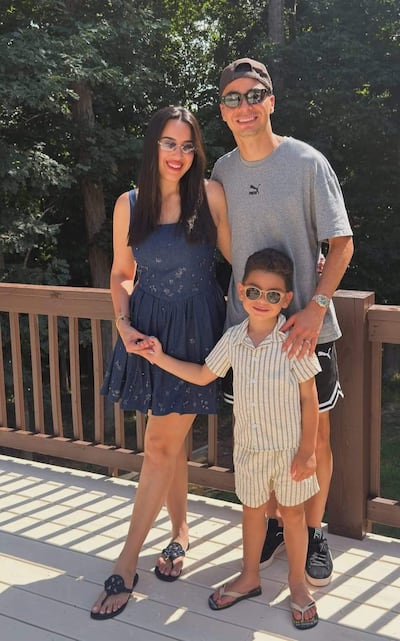 Una mujer en vestido azul y gafas de sol sonríe con un niño en conjunto a rayas, mientras un hombre con camiseta gris la abraza.