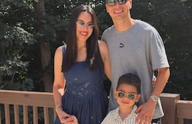 Una mujer en vestido azul y gafas de sol sonríe con un niño en conjunto a rayas, mientras un hombre con camiseta gris la abraza.