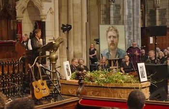 Un hombre de pie junto a un atril con vela, dirigiéndose a una audiencia en un ambiente introspectivo, rodeado de guitarras y un retrato.