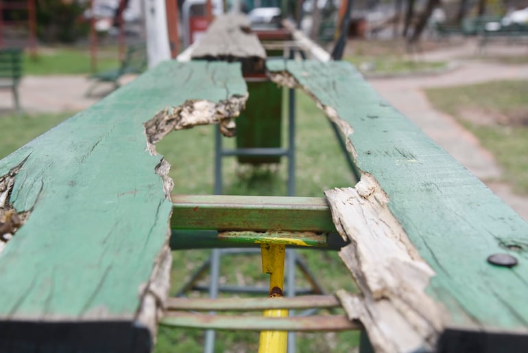 Banco deteriorado de color verde con sección faltante, rodeado de césped en un parque, sin personas presentes.