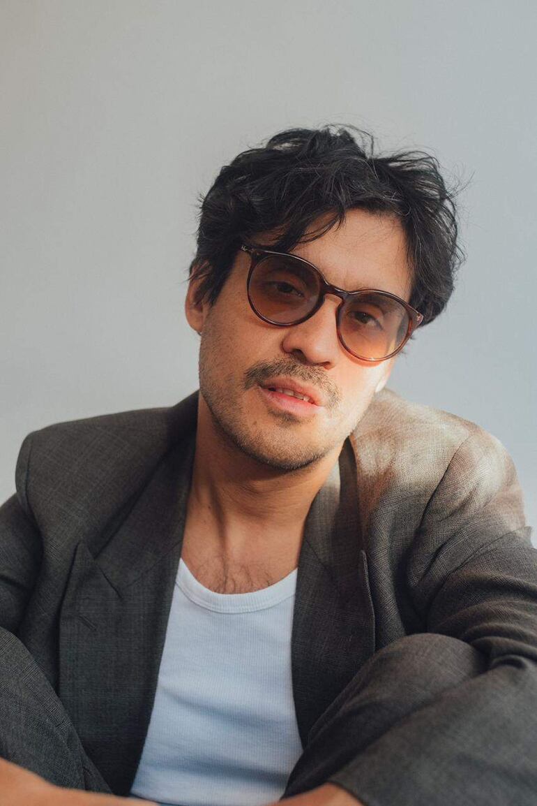 El David Aguilar, con traje gris y gafas de sol grandes, sentado con expresión pensativa en fondo blanco.