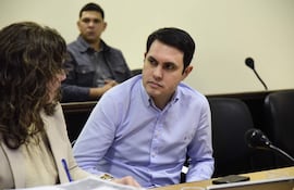 Hombre con camisa de cuadros azules y mujer con cabello rizado en chaqueta clara, conversan frente a un escritorio lleno de documentos.