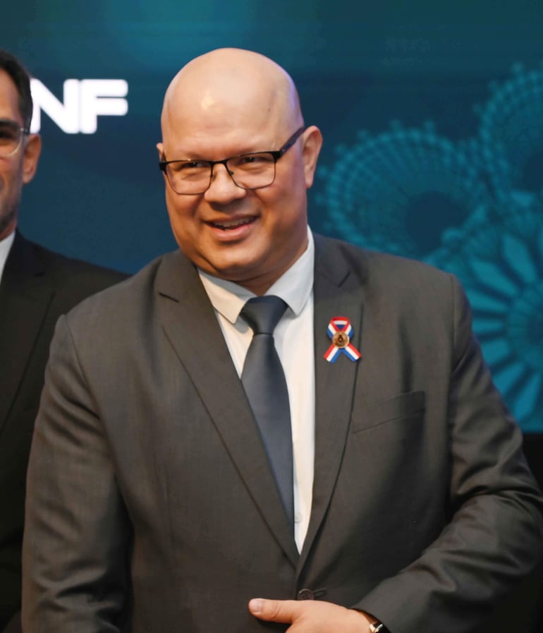 Óscar Lovera, calvo y con gafas, sonríe en traje oscuro con corbata azul y medalla patria; fondo oscuro y otros participantes en penumbra.