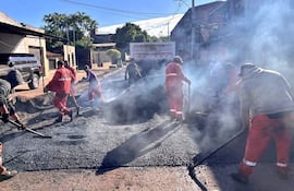 Siete obreros en camisetas rojas y grises manipulando el asfalto en una calle, rodeados de humo y edificios urbanos.