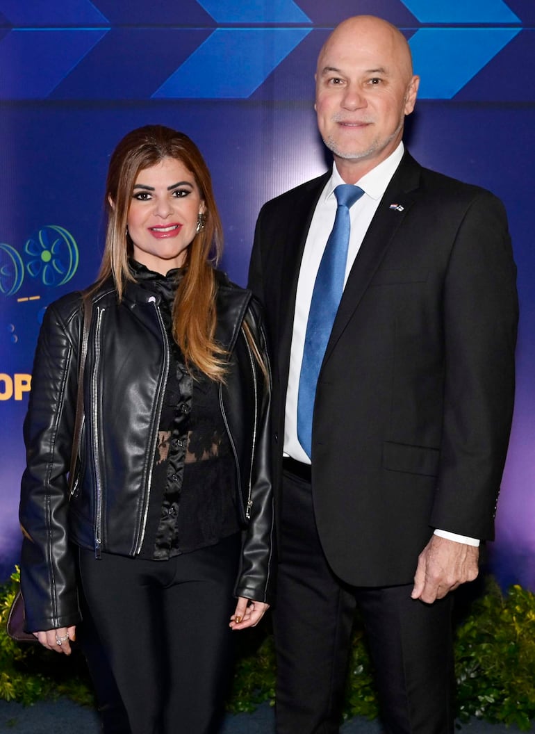 Mujer con chaqueta de cuero negro y hombre en traje oscuro sonríen en un evento formal con fondo azul.