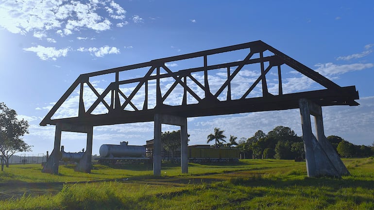 Estructura metálica en un entorno verde, con un cielo parcialmente nublado y restos de instalaciones industriales en el fondo.