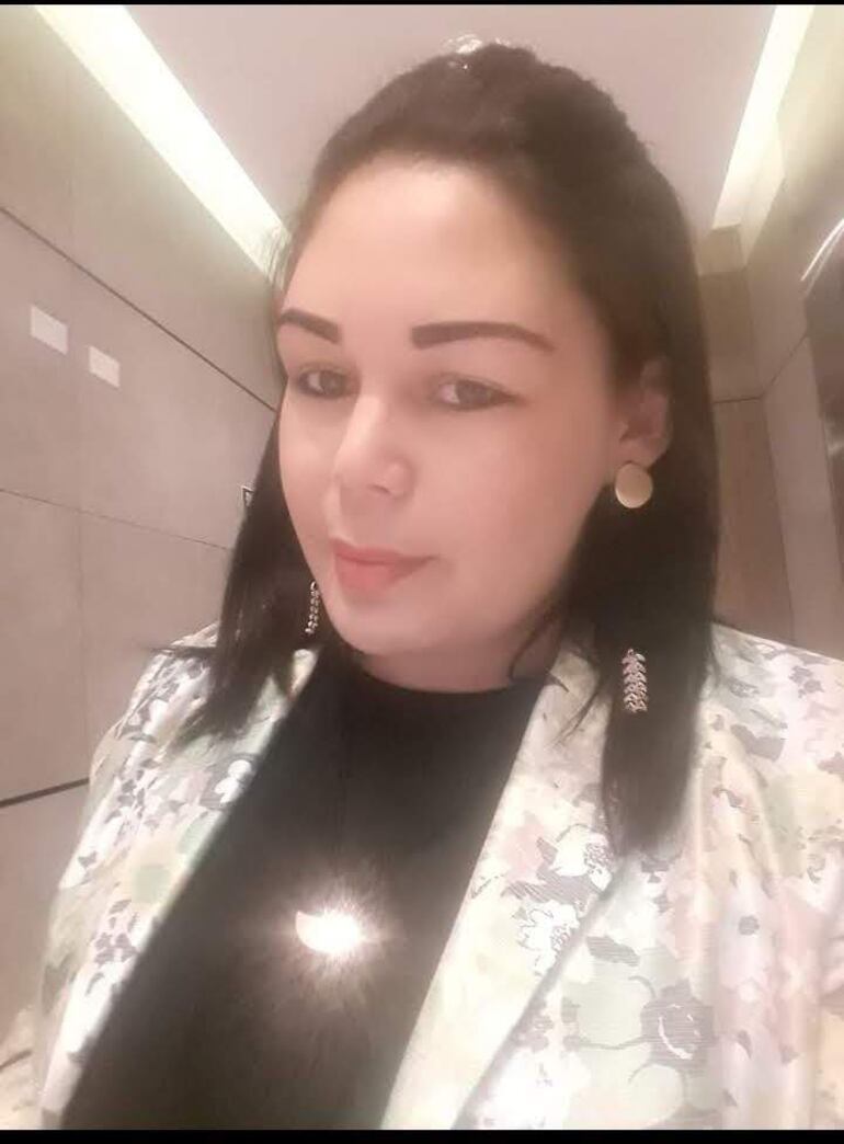Mujer con cabello largo y oscuro, blusa negra y abrigo floral, sonríe en un ambiente iluminado suavemente.
