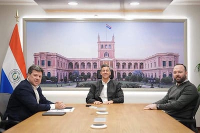 Santiago Peña sonríe con chaqueta negra, flanqueado por Rolando De Barros en suéter gris y otro hombre en camisa blanca, en un ambiente formal.