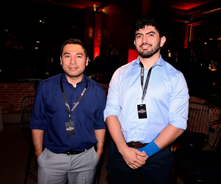 Dos hombres sonrientes en camisa, uno azul y otro claro, con credenciales en un ambiente cálido y decorativo.