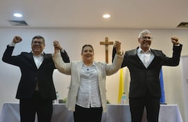 Tres individuos en celebración: mujer con blusa blanca y saco claro, hombres con trajes oscuros, fondo con cruz.